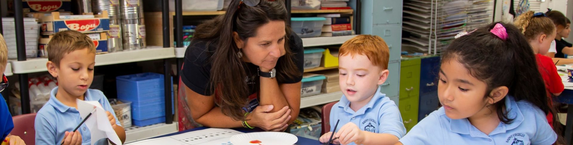 young male and female children in light blue uniform shirts painting at a blue table with a female teacher leaning over the table to assist