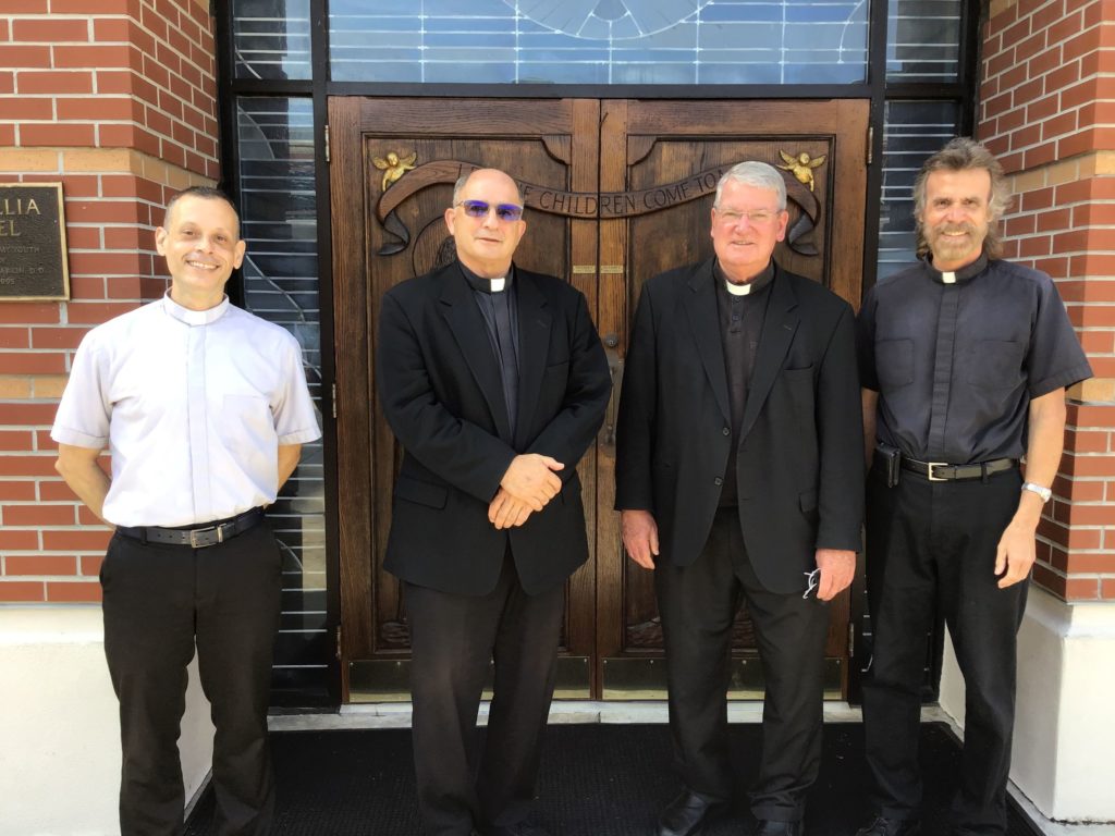 four priests dressed in black and white in front of the the doors of st cecelia catholic school